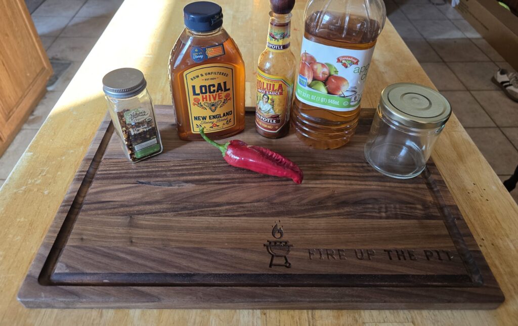 Hot honey ingredients on a Fire Up The Pit cutting board: honey, crushed red pepper, chipotle hot sauce, apple cider vinegar, a red cowhorn pepper and a glass jar