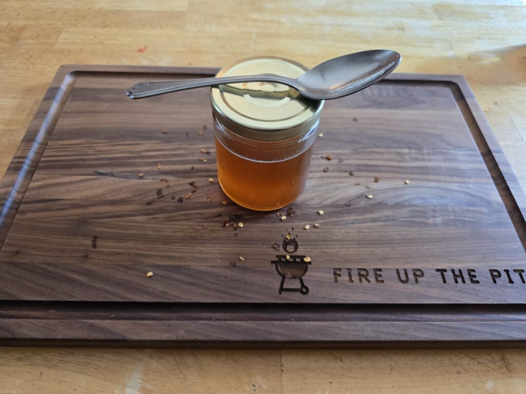 Jar of homemade hot honey with a spoon on the lid and red pepper flakes on a Fire Up The Pit cutting board