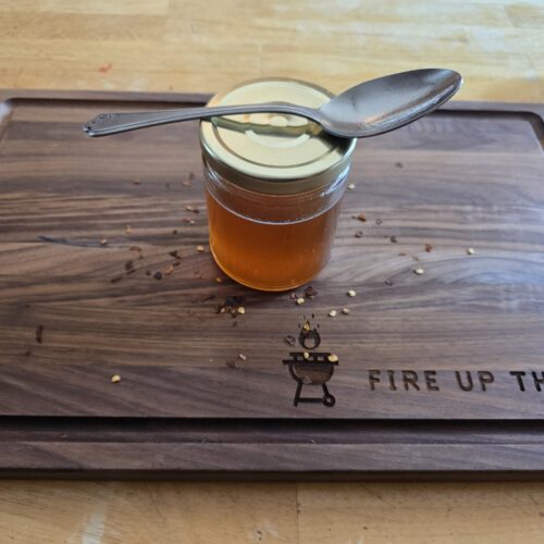 Jar of homemade hot honey with a spoon on the lid and red pepper flakes on a Fire Up The Pit cutting board